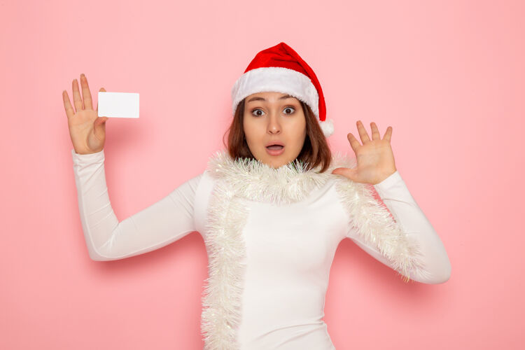 肖像正面图年轻女性手持白色银行卡在粉色墙壁上圣诞节彩色节日新年时尚帽子银行色彩