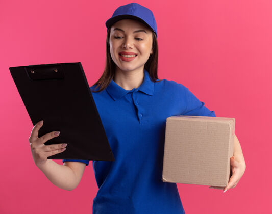 保持穿着制服的微笑漂亮的女送货员拿着名片盒 看着隔离在粉色墙上有复印空间的剪贴板女人送货员微笑