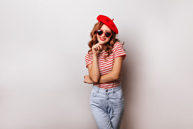 深色快乐的法国女孩穿着牛仔裤表达积极的情绪美丽的女模特戴贝雷帽和太阳镜在白墙上微笑头发帽子波浪发