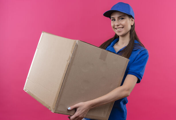 制服身穿蓝色制服 头戴帽子的年轻送货女孩拿着大盒子 面带微笑地看着相机包装盒子脸