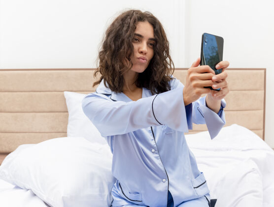 漂亮穿着蓝色睡衣的年轻美女坐在床上用智能手机自拍 在卧室内部灯光背景下吹一吻智能手机吹女人