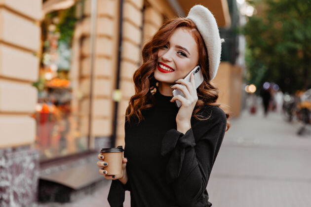 年轻快乐的欧洲女孩戴着贝雷帽微笑着站在城墙上迷人的长发女士一边打电话一边喝咖啡街头风格微笑红发