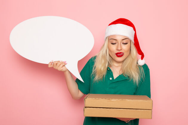 圣诞节正面图年轻女性手持白色大牌子和食品盒在粉色墙上拍照工作新年假期工作快递制服风景盒子工作