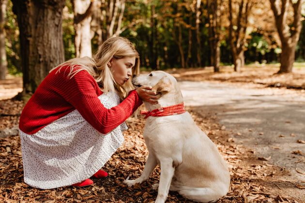朋友美丽的女人亲吻她可爱的漂亮的狗穿着红色毛衣和白色裙子的可爱女孩与宠物分享爱成人玩耍户外