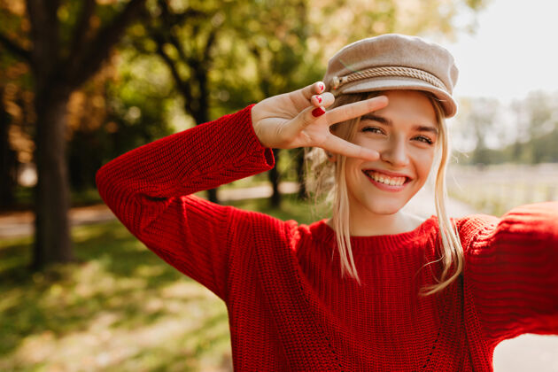 多漂亮的金发女孩戴着时髦的帽子 穿着红色毛衣 微笑着在公园里自拍光季节户外
