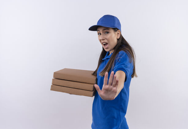 表情身穿蓝色制服 头戴鸭舌帽的年轻送货女孩手拿比萨饼盒 脸上带着恐惧的表情 在做停车标志做标志女孩