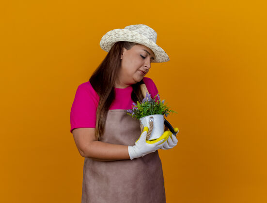 女人穿着围裙戴着帽子的中年园丁妇女拿着铲子和盆栽植物站在橙色的背景上面带微笑地看着植物看中间围裙