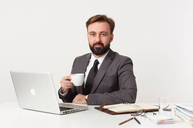 胡须照片中 年轻的留着胡须的黑发男性留着短发 面色平静地看着前方 一边用笔记本电脑和笔记本在白墙上工作 一边喝茶短嘴唇肖像