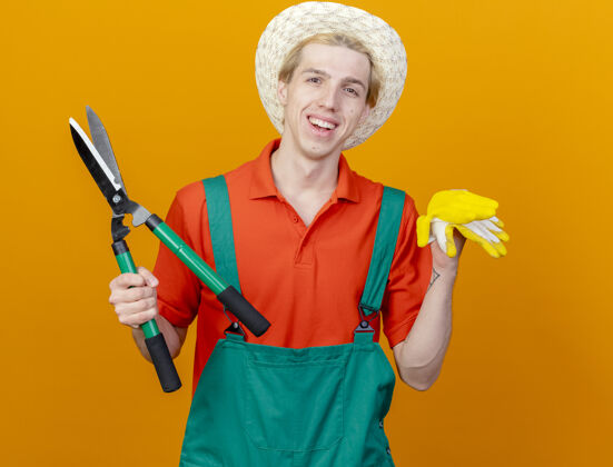 手套年轻的园丁穿着连体衣 戴着帽子 拿着树篱剪和橡胶手套 站在橙色的背景上开心地微笑着看着镜头站帽子穿