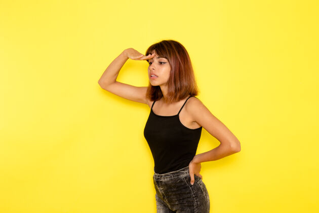 年轻身穿黑色衬衫的年轻女性在浅黄色墙上遥望远方的前视图性感情感模特