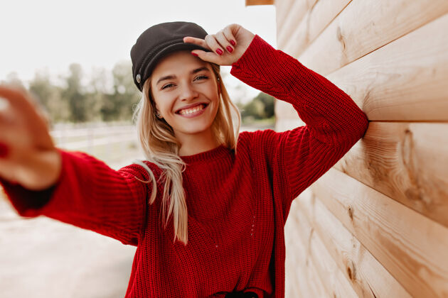 快乐穿着红色套头衫 戴着黑帽子的迷人金发女郎开心地对着手机微笑漂亮女孩在户外木屋附近自拍微笑房子户外