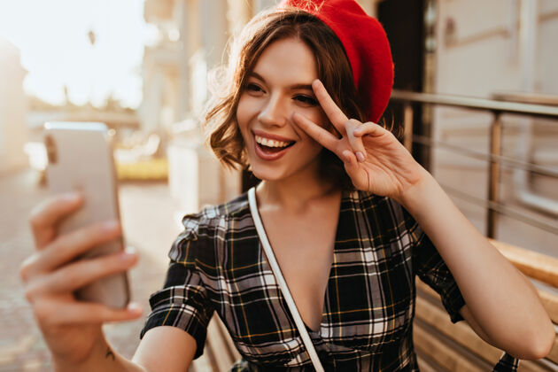 积极穿着红色贝雷帽的兴奋女孩在街上用智能手机自拍可爱的白种女人在拍照 看着手机屏幕城镇成人电话