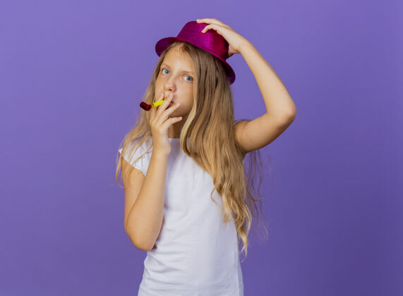 派对美丽的小女孩在节日帽子吹哨子快乐和积极 生日聚会的概念站在紫色的背景吹漂亮女孩