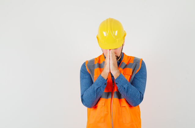 肖像年轻的建筑工人穿着衬衫 背心 头盔 手举祈祷的姿势 神情平静 俯瞰前方手冷静工人
