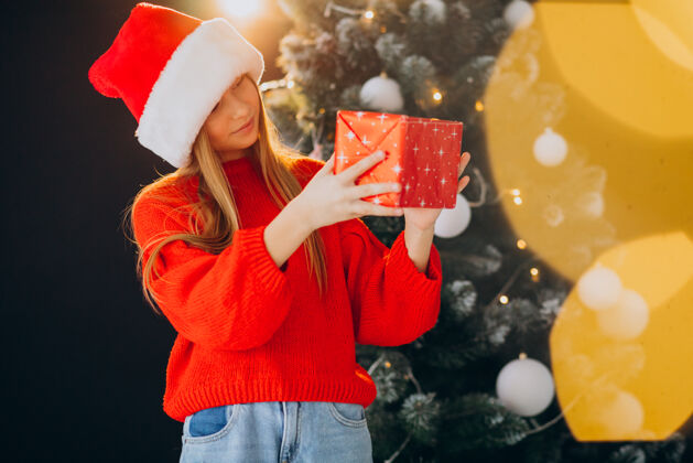 看可爱的少女戴着圣诞树旁的红色圣诞帽年轻房子灯