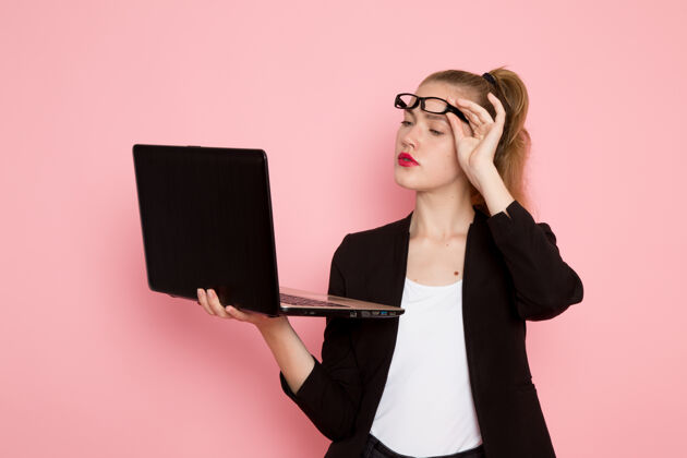 商务穿着黑色夹克的女上班族在浅粉色墙上使用笔记本电脑的前视图时尚人办公室