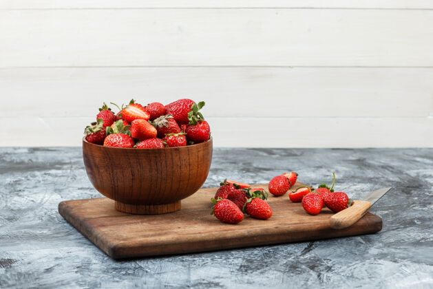 特写一碗草莓和一把刀放在深蓝色大理石和白色木板背景上的木质砧板上特写为您的文字腾出空间刀美味浆果