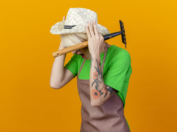 表情一位年轻的园丁 围裙上留着短发 戴着帽子 手里拿着小耙子 站在橙色的背景上 脸上露出恼怒的表情年轻围裙抱