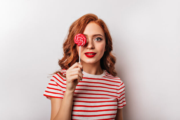 积极可爱的红发女孩拿着棒棒糖在白墙上摆姿势迷人的年轻女子拿着红糖年轻糖果头发
