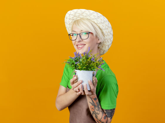 看一位年轻的园丁 围裙上戴着短发 戴着帽子 展示着盆栽植物 站在橙色的背景下 面带微笑地看着摄像机花园脸立场
