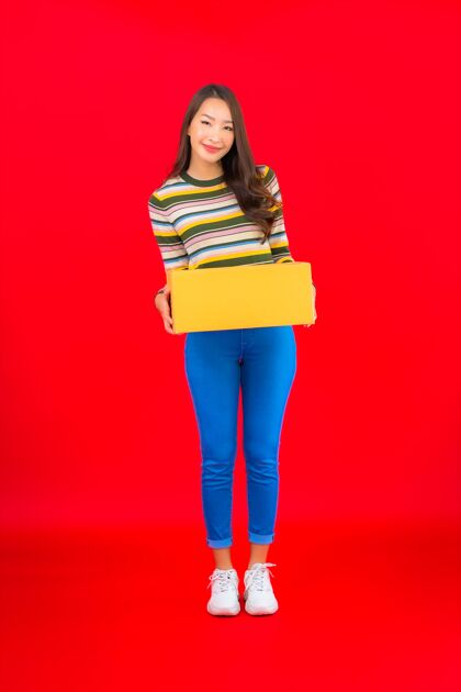交货在红色的隔离墙上画一幅美丽的亚洲年轻女子拿着包裹盒商业购物企业家