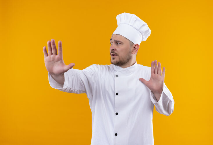 伸展年轻帅气的厨师身着厨师制服 伸出手看着橙色空间上孤立的一面厨师壁板手
