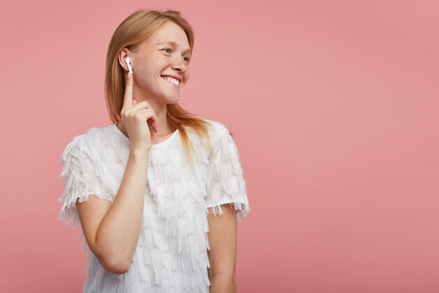 T恤活泼迷人的年轻女士 一头狐狸精的头发 把听筒插进耳朵里 面带幸福的笑容 积极地看着一边 穿着白色优雅的t恤 站在粉色的背景上休闲耳机手