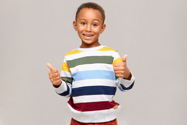 微笑人类积极的情绪 反应和感受情绪化快乐的深色皮肤男孩 穿着多色套头衫 竖起大拇指 表示同意 赞同 表达自己的喜欢 面带笑容童年拇指孩子