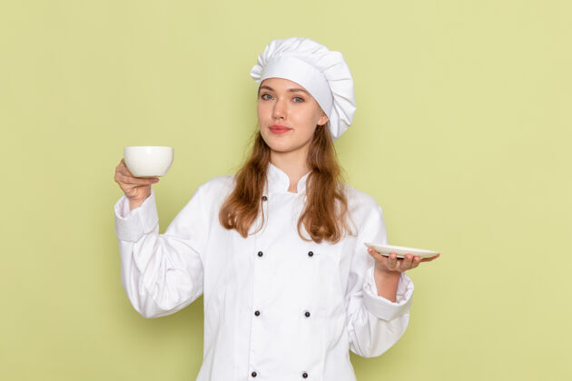 举行身着白色厨师服的女厨师正对着绿色墙上的杯子和盘子烹饪帽子盘子