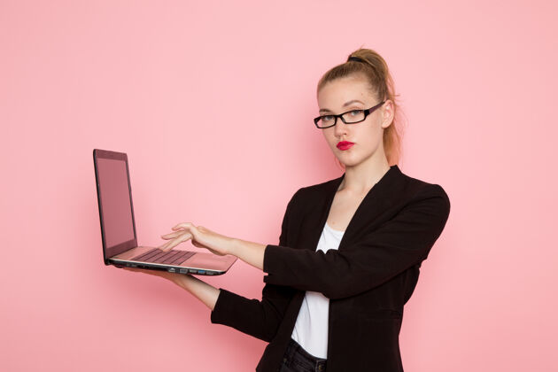 严格穿着黑色夹克的女上班族在浅粉色墙上使用笔记本电脑的前视图个人公司笔记本电脑