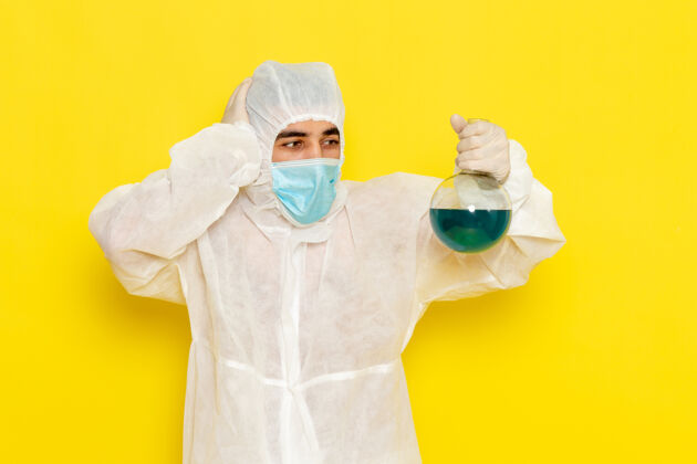 科学穿着特殊防护服的男科学工作者的正面图 浅黄色的墙上有带面罩的瓶子烧瓶防护手持