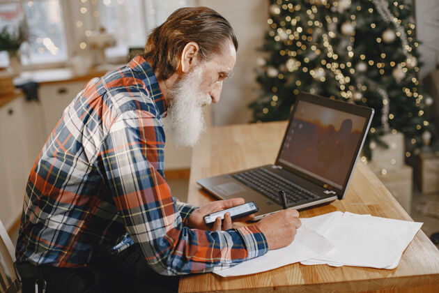工作老人拿着笔记本电脑爷爷坐在圣诞装饰品里老人拿着手机胡须笔记本电脑成人
