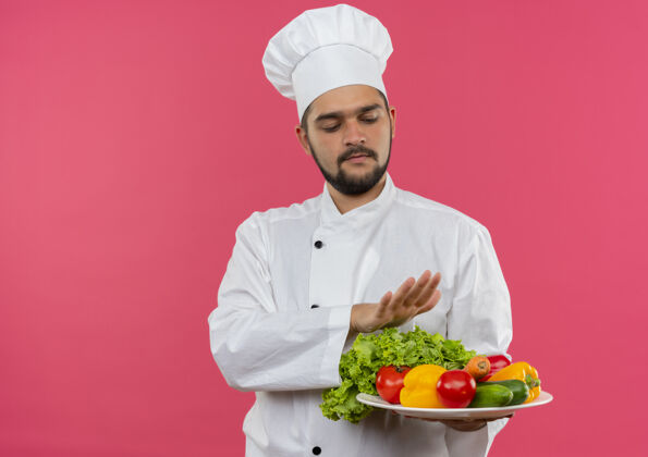 年轻年轻的男厨师穿着厨师制服 拿着和看着盘子里的蔬菜 把手放在盘子上面 把粉红色的空间隔离开来保持盘子粉红