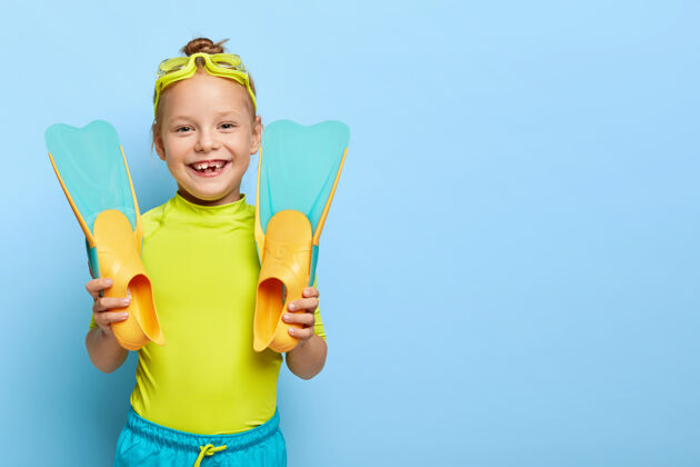 人快乐姜小女孩的照片展示了新的橡胶脚蹼 戴着泳镜 穿着夏装 喜欢学习游泳 有积极的休息 隔离在蓝色的墙上空白年轻泳装红发