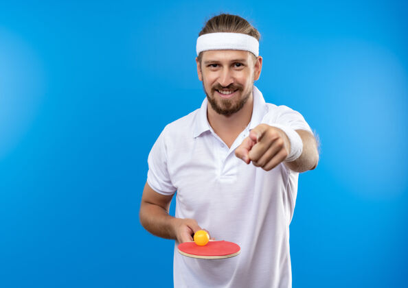 空间面带微笑的年轻帅气的运动型男人戴着头带和腕带拿着乒乓球拍拿着球孤立地指着蓝色的空间球拍乒乓球英俊