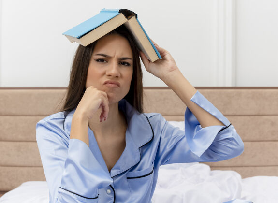 相机穿着蓝色睡衣的年轻漂亮女人坐在床上 头上举着书 看着卧室里灯光背景下的相机 不高兴漂亮睡衣看着
