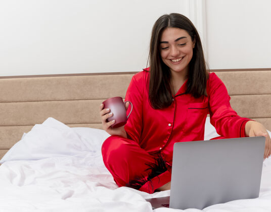 咖啡穿着红色睡衣的年轻美女坐在床上 手里拿着笔记本电脑和一杯咖啡 在卧室的室内灯光背景下愉快地微笑着卧室快乐女士