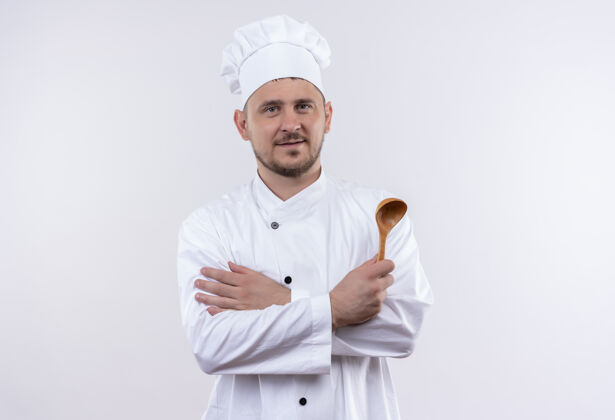 年轻高兴的年轻帅哥厨师穿着厨师制服拿着勺子站着封闭的姿势隔离在空白处烹饪制服请