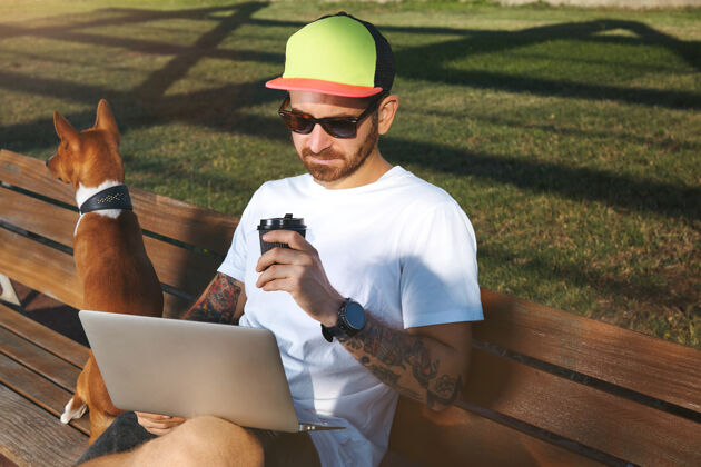 肖像一个留着胡须和纹身的年轻人穿着一件纯白的t恤 一边喝着咖啡 一边看着他的笔记本电脑 而他的棕色和白色的狗坐在公园的长凳上休闲城市早晨