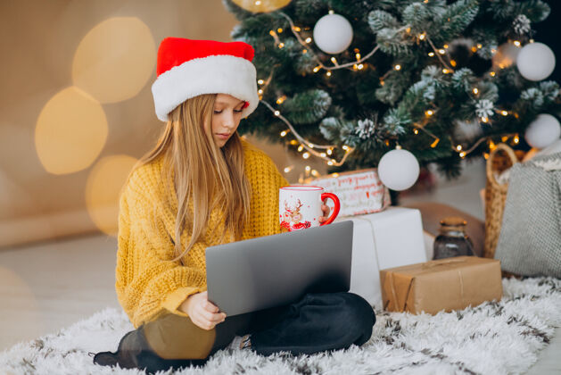 红帽子可爱的女孩在圣诞树旁用电脑帽子视频通话销售
