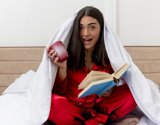 女人穿着红色睡衣的年轻美女坐在床上 用毯子裹着一杯咖啡和一本书 愉快而积极地微笑着 在卧室内部的灯光背景下快乐睡衣书