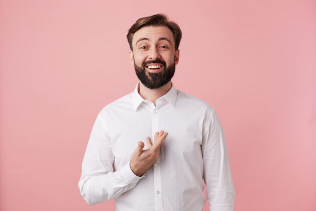 成人摄影棚照片中 年轻漂亮的留着胡须的黑发男性面带微笑 愉快地站在前面 身着正式服装 在粉色的墙上摆姿势 露出洁白完美的牙齿积极发型美丽