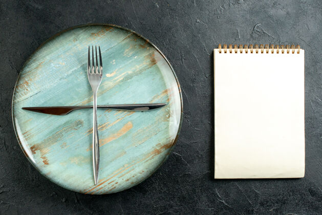 时针顶视图交叉叉子和刀子在青色圆板笔记本在黑暗的桌子上手时间模拟