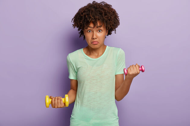 私人教练迷惑不解的非洲裔美国妇女看起来很惊讶 拿着两个哑铃 锻炼肌肉 穿t恤 喜欢健身和运动 孤立在紫色的墙上女子举重训练在健身房休闲爱好长相