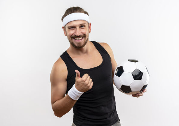 展示面带微笑的年轻英俊的运动型男子戴着头带和腕带拿着足球 在空白处孤立地竖起大拇指向上球英俊