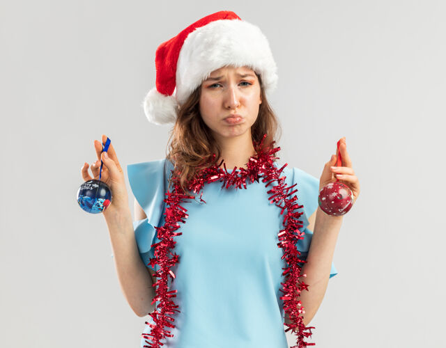 新年心烦意乱的年轻女子身穿蓝色上衣 戴着圣诞帽 脖子上戴着金属丝 手里拿着圣诞球 表情悲伤顶女人表情