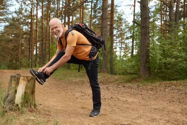 散步户外夏季拍摄健康的老年男性 背着背包 脚放在树桩上 在森林里摆姿势 在运动鞋上系鞋带 准备长时间攀爬 带着快乐的微笑远足男人健身鞋带