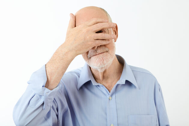 成人穿着蓝色衬衫的高加索退休老人的肖像 手放在脸上 遮住眼睛 从裂开的手指中向外窥视 感到羞愧人类的面部表情和肢体语言英俊隐藏表情