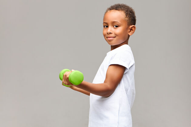 力量一个英俊快乐的非洲男孩用健身器材在灰色的空白墙上摆姿势 举着绿色的哑铃 锻炼手臂肌肉 面带幸福的微笑活跃非洲运动