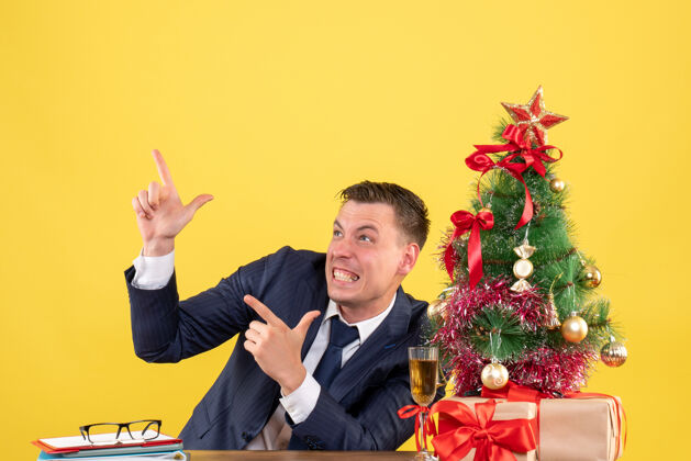 人前视图兴高采烈的男子坐在桌旁手指着圣诞树附近的东西和黄色的礼物树兴高采烈西装